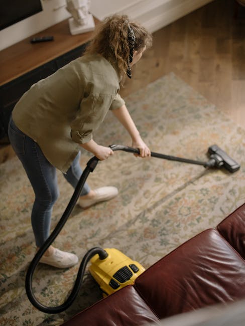 A person using a yellow vacuum cleaner to perform deep surface cleaning on an ornate patterned rug in a living room, with a wooden floor and a brown leather sofa in the background. The room is well-li
