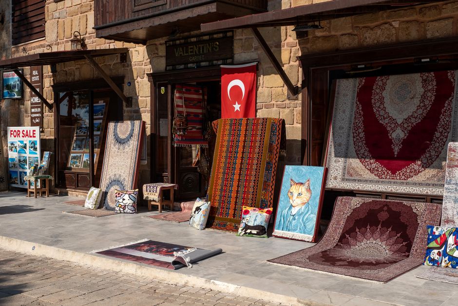 A black-and-white photograph of a building facade with a sign reading 'Gülzar Carpet,' featuring two large windows with wooden shutters and framed artwork displayed outside on the wall. Several tradi